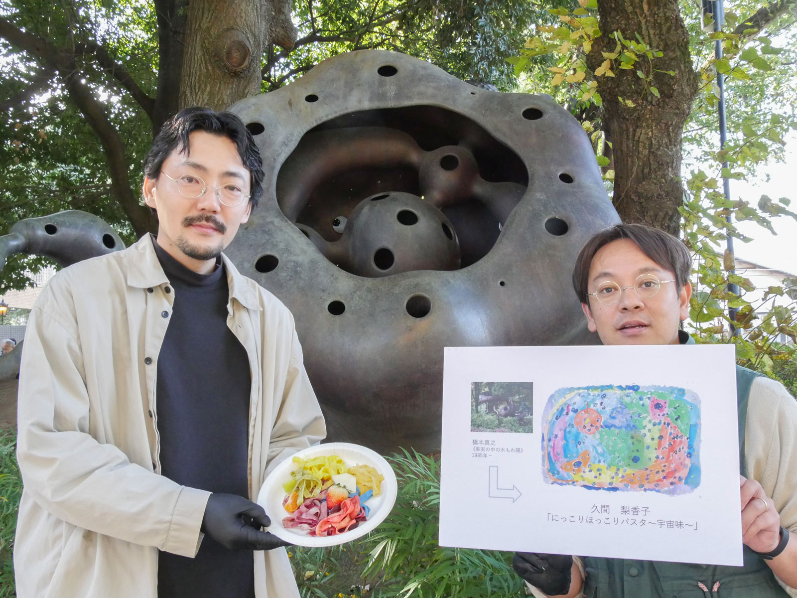 課題作品となった屋外彫刻の前で、総合グランプリ受賞作品パネルと創作したパスタを持つL PACK.の2人